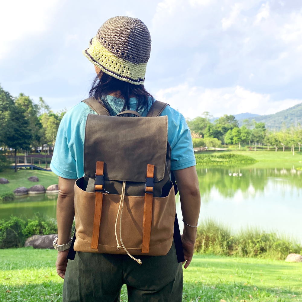 Person with a brown backpack and knit hat standing by a lake with greenery and mountains in the background