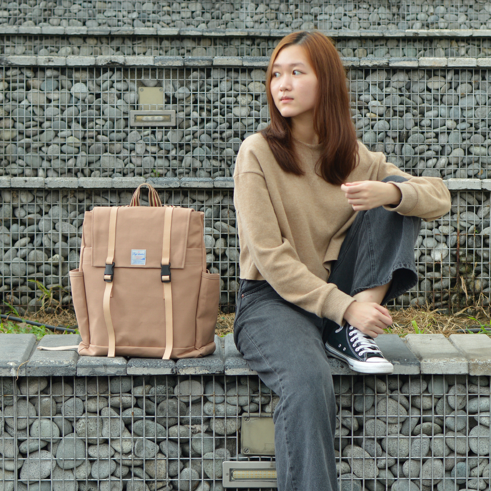 Woman sitting on a stone wall with a beige backpack next to her against a stone wall background