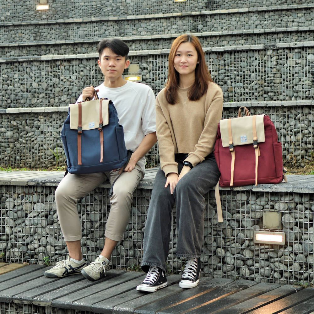 Two people sitting on a wooden bench with two bags, one blue and one red.